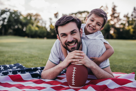 Handsome dad with his little cute sun are laying on green grassy lawn on American flag with American football ball in handの写真素材