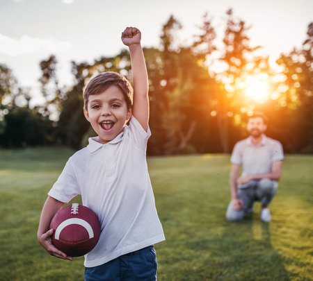 Handsome dad with his little cute sun are having fun and playing American football on green grassy lawnの写真素材