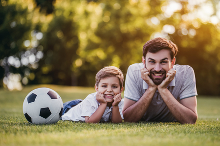 Handsome dad with his little cute sun are having fun and playing football on green grassy lawnの写真素材