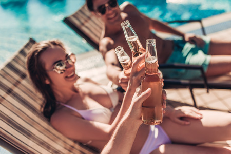 Cheers! Group of young friends is lying on a chaise-longues near swimming pool and smiling. Having rest and drinking beer together.の写真素材