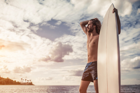 Young handsome man with long hair is standing on beach with white surfboard in hands.