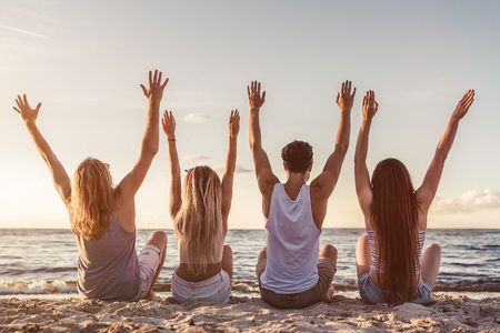 Back view of group of young attractive friends are sitting on beach with raised hands up.の写真素材