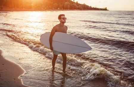 Young handsome sporty man is walking on beach with white surfboard in hands.の写真素材