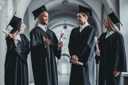 Happy graduates are standing in university hall in mantles with diplomas in hand.の写真素材