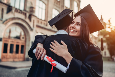 Graduates in mantles with diplomas in hands are standing near university and hugging each other.の写真素材