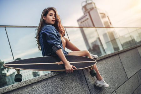 Young woman is posing with skateboard in the city. Female teenager outdoor with longboard.