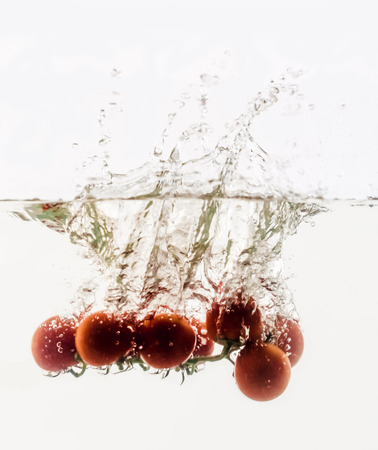 Vegetables are thrown into the water in transparent vessel. Tomatoes on a branch and water splash on white background.の写真素材