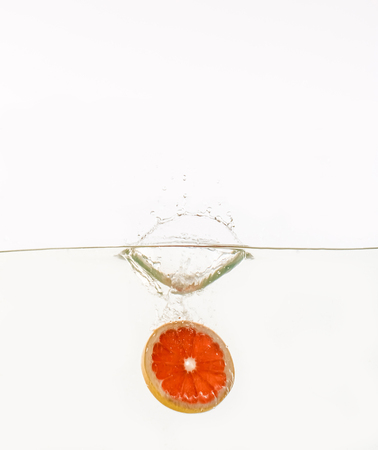 Cut fruits are thrown into the water in transparent vessel. Grapefruit and water splash on white background.の写真素材