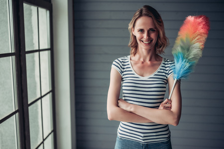 Beautiful young woman is doing cleaning at home. Holding cleaning tools, looking at camera and smilingの写真素材