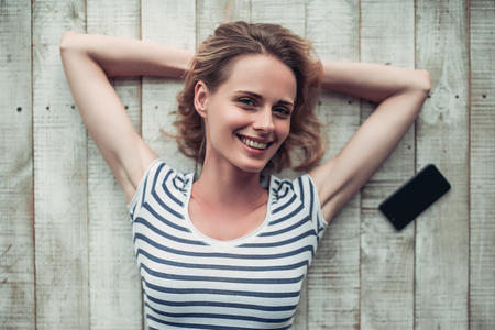 Young beautiful woman is lying on the floor with mobile phone nearby. Smiling and looking at the camera.の写真素材