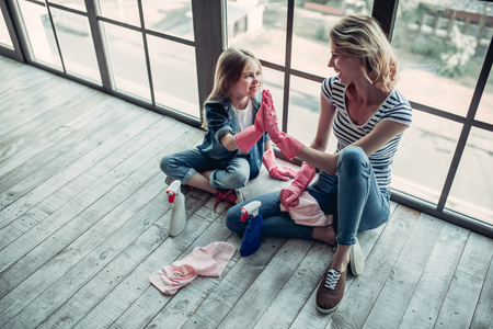 Beautiful young woman  and her little cheerful daughter are doing cleaning at home. Sitting near the cleaning tools and smiling. Good job! Giving five to each other.の写真素材