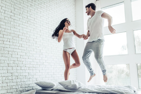 Happy young couple is having fun in bedroom. Enjoying the company of each other while jumping on bedの写真素材