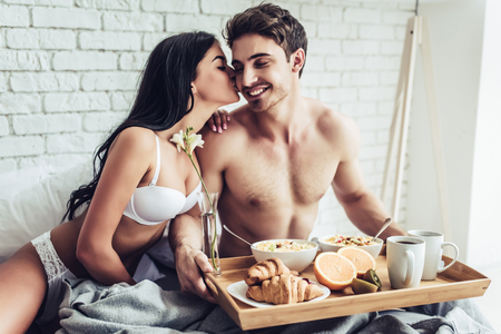 Good morning! Handsome man brings healthy breakfast in bed. Young beautiful love couple is sitting in bed before having breakfast.の写真素材