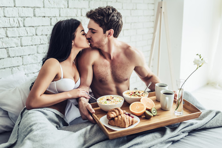 Good morning! Handsome man brings healthy breakfast in bed. Young beautiful love couple is sitting in bed before having breakfast.の写真素材