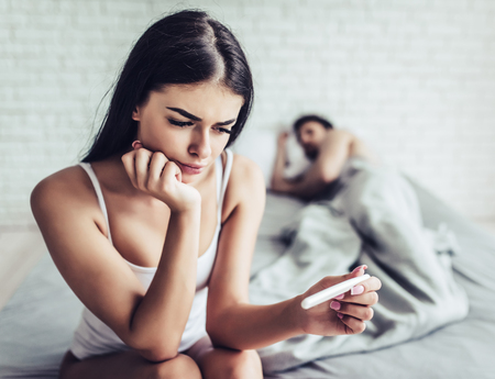 Young pensive woman is sitting on the edge of bed with pregnancy test in hand. Man is sleeping on the background.の写真素材