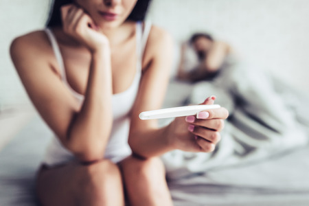 Cropped image of young woman is sitting on the edge of bed with pregnancy test in hand. Man is sleeping on the background.の写真素材
