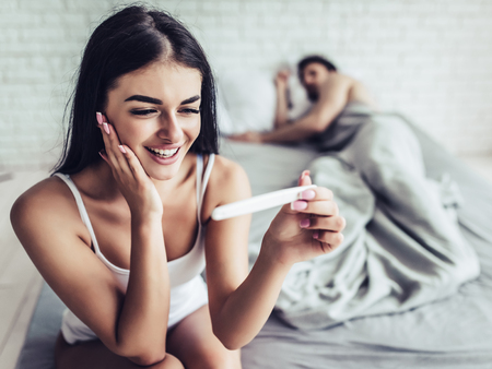 Young happy woman is sitting on the edge of bed with pregnancy test in hand. Man is sleeping on the background.の写真素材