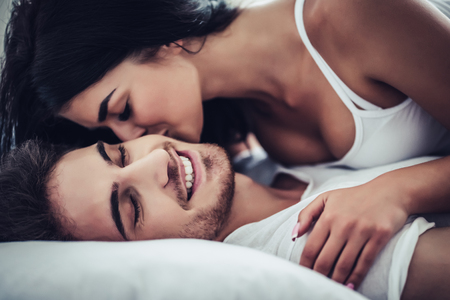 Happy couple is lying in bed together. Enjoying the company of each other. Young woman is kissing her handsome man.の写真素材