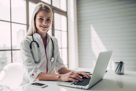 Attractive young female doctor is working with laptop in light cabinet. Smiling and looking at the camera.の写真素材