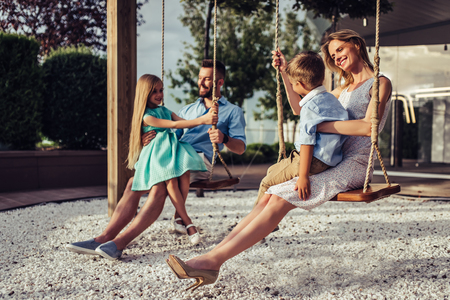 Beautiful family is having fun outside. Parents with children riding on a swing. Mom with dad are playing with their little daughter and son on a terrace with swings.の写真素材