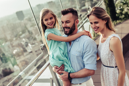 Beautiful family is spending time together  outside. Parents with their little daughter on a roof terrace with view on a city. Mom and dad are having fun with their charming daughter.の写真素材