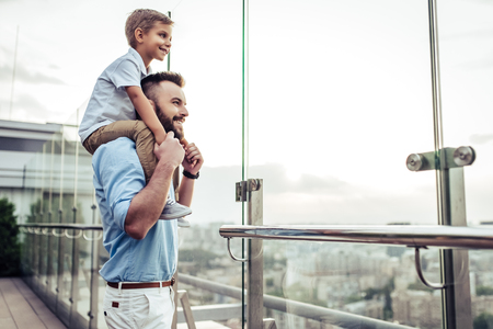 Beautiful family is spending time together  outside. Dad and his little son are having fun on a roof terrace with view on a city. Sitting on father's shoulders and smiling.の写真素材