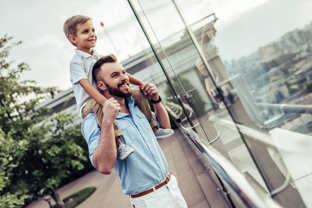 Beautiful family is spending time together  outside. Dad and his little son are having fun on a roof terrace with view on a city. Sitting on father's shoulders and smiling.の写真素材