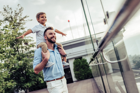 Beautiful family is spending time together  outside. Dad and his little son are having fun on a roof terrace with view on a city. Sitting on father's shoulders and smiling.の写真素材