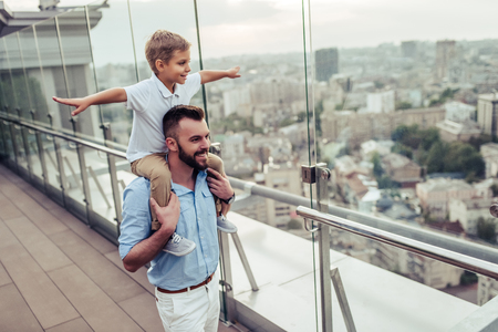 Beautiful family is spending time together  outside. Dad and his little son are having fun on a roof terrace with view on a city. Sitting on father's shoulders and smiling.の写真素材