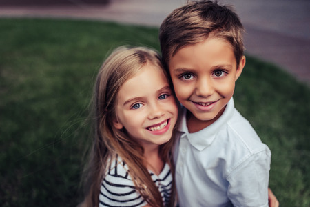 Couple of charming childs having fun outdoors. Portrait of beautiful girl and boy are hugging, smiling and looking at the camera. Little brother and sister in park on a green grass.の写真素材