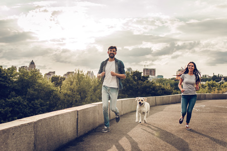 Romantic couple is on a walk in the city with their dog labrador. Beautiful young woman and handsome man are running and having fun outdoors with golden retriever labrador.の写真素材