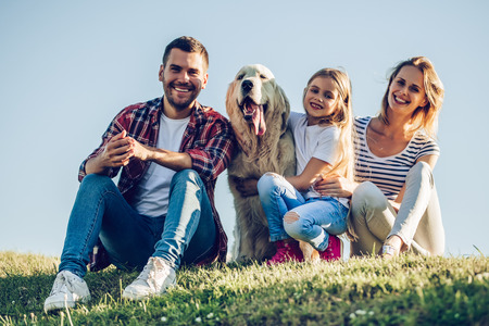 Beautiful happy family is having fun with golden retriever outdoors. Mother, father and daughter are sitting with dog labrador on green grass in park.の写真素材