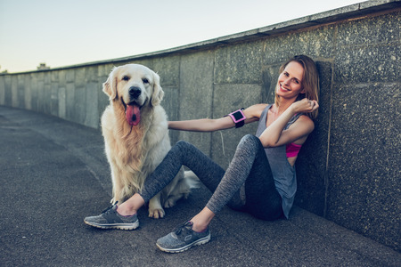 Attractive sporty woman in the city with dog golden retriever during running. Taking break, listening to music and smiling.の写真素材