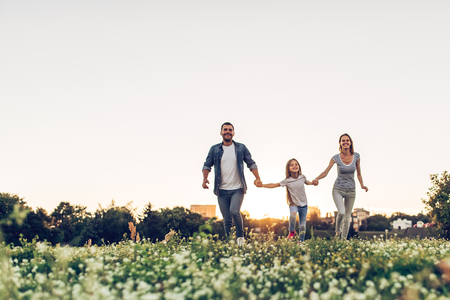 Happy family outdoors spending time together. Father, mother and daughter are having fun and running on a green floral grass during the sunset.の写真素材