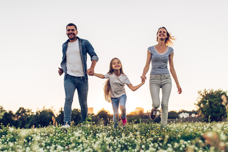 Happy family outdoors spending time together. Father, mother and daughter are having fun and running on a green floral grass during the sunset.の写真素材