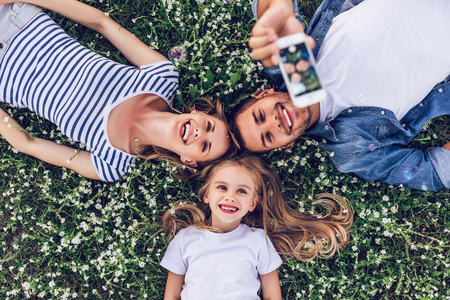 Top view of happy family outdoors spending time together. Father, mother and daughter are having fun and taking selfie on a smart phone while lying on a green floral grass.の写真素材