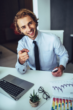 Hey you Young handsome businessman is working in light modern office with laptop, smiling and looking at the camera.の写真素材