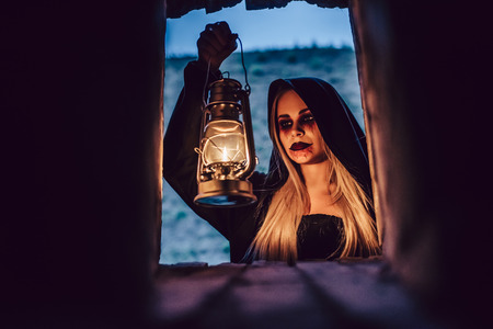 Happy Halloween! Portrait of black witch with scary makeup in black mantle with lamp in hands.の写真素材