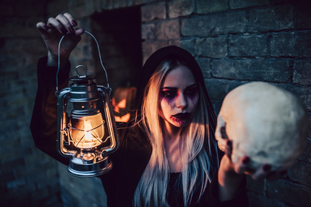 Happy Halloween! Portrait of black witch with scary makeup in black mantle with lamp and skull in hands. Pumpkins are on the background.の写真素材