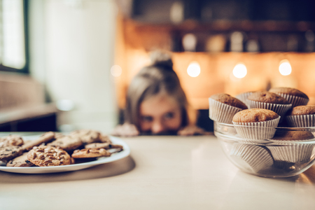 Charming little girl on kitchen is looking from under the table on sweets. Ready to eat some cookies and cakes.の写真素材