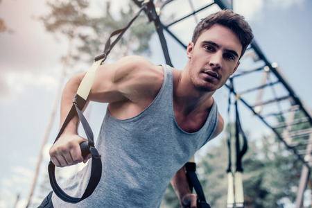 Handsome muscular man is training outdoor on TRX. Total Body Resistance Exercises. Working out at...
