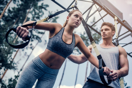 Young couple are training outdoor on TRX. Total Body Resistance Exercises. Attractive woman is...