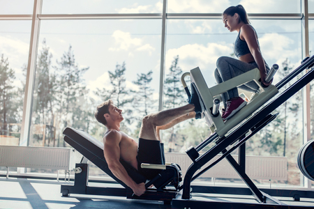 Young couple is working out at gym. Attractive woman and handsome muscular man are training in light modern gym. Man is doing leg press.の写真素材