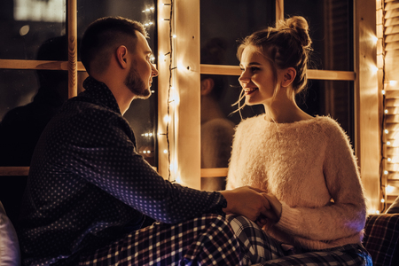 Young romantic couple is spending time together at home in pajamas.Sitting on window sill with garland in New Year Eve.Waiting for Christmas.Two lovers are hugging and kissing in Saint Valentine's Dayの写真素材