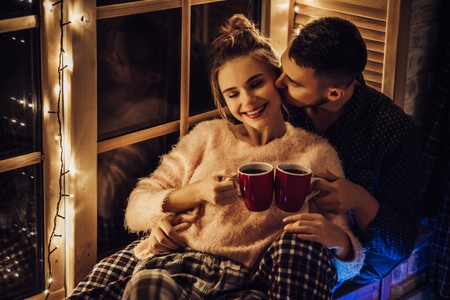 Young romantic couple is spending time together at home in pajamas.Sitting on window sill with garland in New Year Eve.Waiting for Christmas.Two lovers are hugging and kissing in Saint Valentine's Dayの写真素材