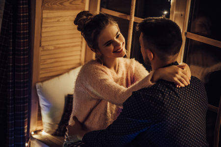 Young romantic couple is spending time together at home in pajamas.Sitting on window sill with garland in New Year Eve.Waiting for Christmas.Two lovers are hugging and kissing in Saint Valentine's Dayの写真素材