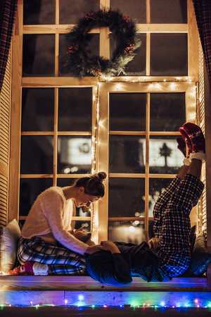Young romantic couple is spending time together at home in pajamas.Sitting on window sill with garland in New Year Eve.Waiting for Christmas.Two lovers are hugging and kissing in Saint Valentine's Dayの写真素材