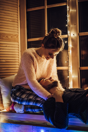 Young romantic couple is spending time together at home in pajamas.Sitting on window sill with garland in New Year Eve.Waiting for Christmas.Two lovers are hugging and kissing in Saint Valentine's Dayの写真素材