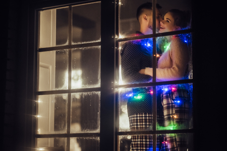 Young romantic couple is spending time together at home in pajamas. Standing near window with garland in New Year Eve. Waiting for Christmas.Two lovers are hugging and kissing in Saint Valentine's Dayの写真素材