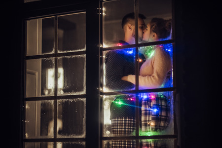 Young romantic couple is spending time together at home in pajamas. Standing near window with garland in New Year Eve. Waiting for Christmas.Two lovers are hugging and kissing in Saint Valentine's Dayの写真素材